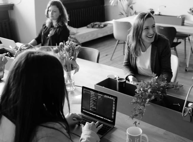 Main image of three females sitting at a desk with one of them with a laptop.